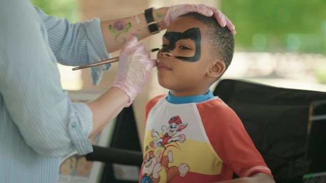 A smiling boy getting a superhero mask painted on his face at an outdoor party