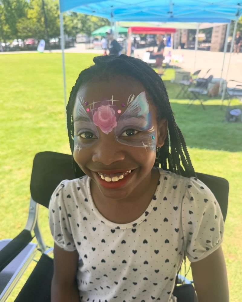A smiling girl with a jeweled butterfly painted around her eyes at an outdoor event