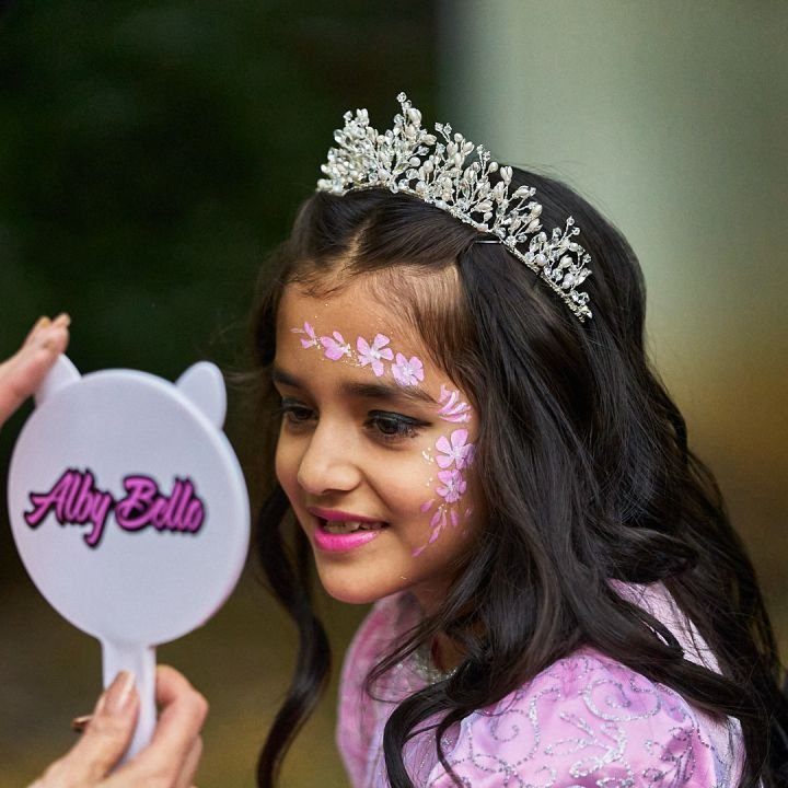 A girl in a sparkly tiara smiling at her floral face painting in a mirror