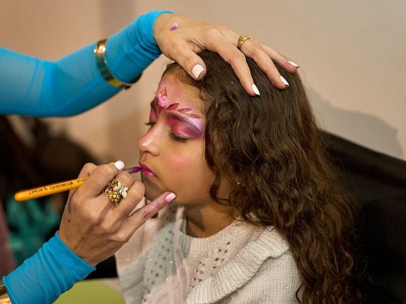 A face painter adding pink and purple swirls around a smiling girl’s eye